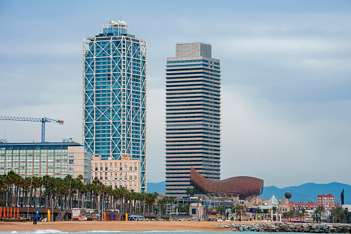 View of Frank Gehry's Golden Fish Sculpture with Mapfre Tower and Hotel Arts at Port Olympic with cloudy sky in the background at Catalonia in Barcelona, Spain