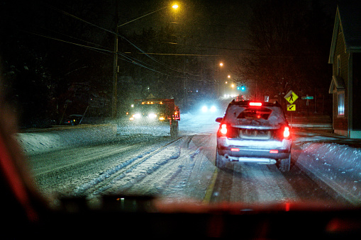 Approaching snowplow dump truck passing stopped traffic at a road intersection during a winter blizzard snow storm viewed through car windshield at night.