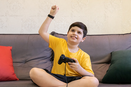 10 year old boy wearing a yellow shirt sitting on the sofa and celebrating an achievement while playing video games.