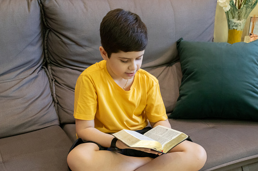 10 year old boy wearing a yellow shirt sitting on the sofa and reading a bible.