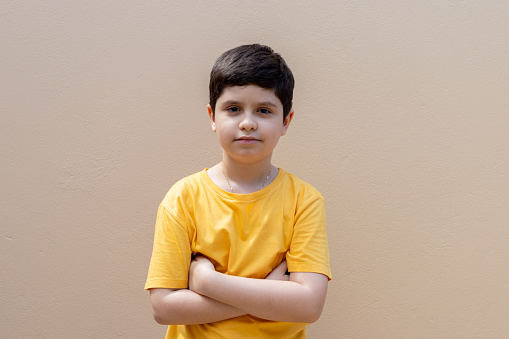 10-year-old boy wearing a yellow shirt with a serious expression and arms crossed against a beige wall in the background.