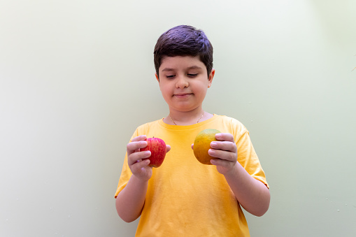 10 year old boy wearing a yellow shirt trying to decide whether to eat an apple or an orange.