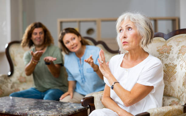 Angry mature female in armchair, keeping distance from the couple sitting on the couch at home Dissatisfied mother-in-law siting on a chair against of a confused husband and wife on the sofa in the living room at home mother in law stock pictures, royalty-free photos & images