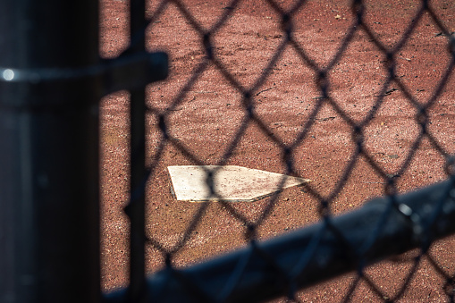 Close up of a white baseball home plate base from behind a black chain link fence in a field of wet brown and red colored dirt or mud on an empty or vacant baseball field on a sunny day.
