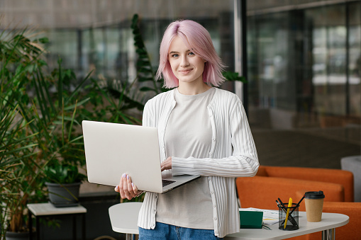 Young successful business woman programmer holding laptop and standing in office or co-working and looking at camera
