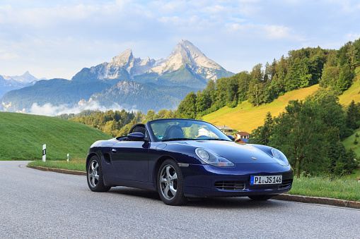 Berchtesgaden, Germany - July 25, 2021: Blue roadster Porsche Boxster 986 with mountain Watzmann and fog panorama. The car is a mid-engine two-seater sports car manufactured by Porsche.