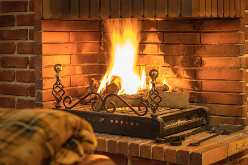 Warming up by the fireplace. Safing energy on heating. Young woman sitting at home by the fireplace and warming her hands, she is wearing white woollen sweater. Cold houses in Europe concept. Energy crisis.