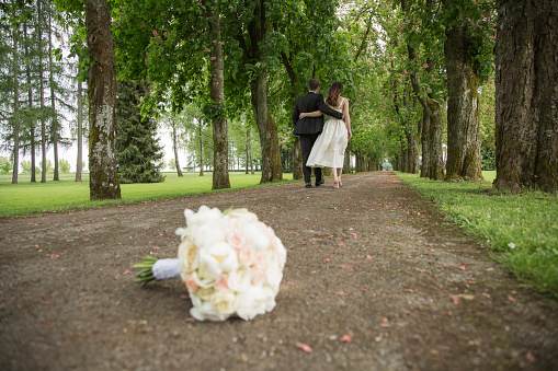 Wedding bouquet of white roses lying on the ground after the wedding.  Bride and groom leaving on treelined path.