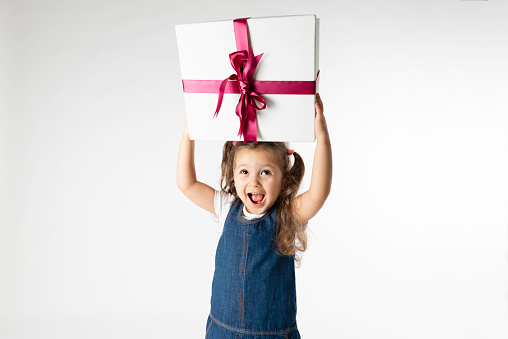 Bright amazed female in white warm knitted outfit and hat holding small wrapped gift excited with Christmas holiday on red backdrop