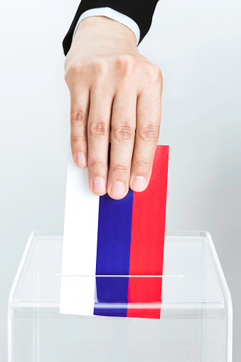 Human hand is inserting russian flag into ballot box.