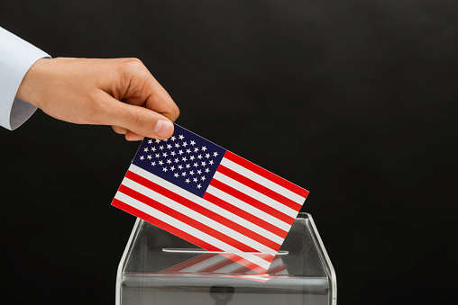 Human hand is inserting American flag into ballot box in front of black background. Representing elections in the United States.