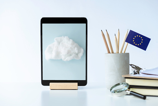 Front view of a white desk with a digital tablet, pen holder with pens and European Union flag, notepad,paperclip, and magnify glass. There is an image of a cloud  on device screen. Representing e-learning in Europe.