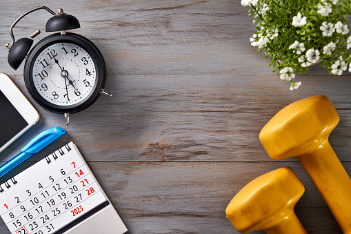 Time to exercise theme: Top view of Clock, calendar, smartphone and dumbbells on a table with copy space