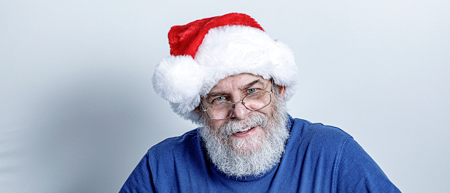 Friendly senior adult man with a curly gray hair full beard is wearing a blue t-shirt and a red and white Santa Claus hat.