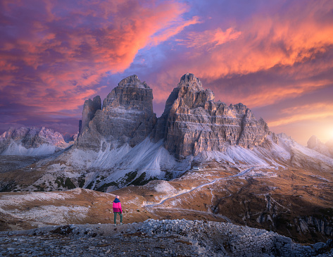 Woman on the hill and mountain peaks at colorful sunset in autumn. Trekking in Dolomites, Italy. Girl on the trail, high rocks at dusk in fall. Landscape with cliffs, purple sky with pink clouds