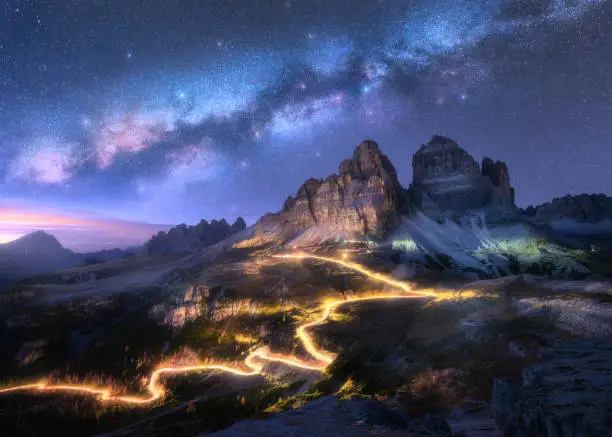 Photo of Milky Way, car light trails on mountain road, high rocks at starry night in summer. Tre cime, Dolomites, Italy. Colorful landscape with blurred light trails, hills, mountain peaks, sky with stars