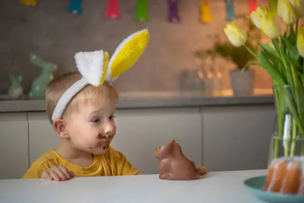Photo of A cute little boy wearing bunny ears on Easter day is eating a chocolate Easter bunny. A child plays egg hunt for Easter. Charming child celebrates Easter at home