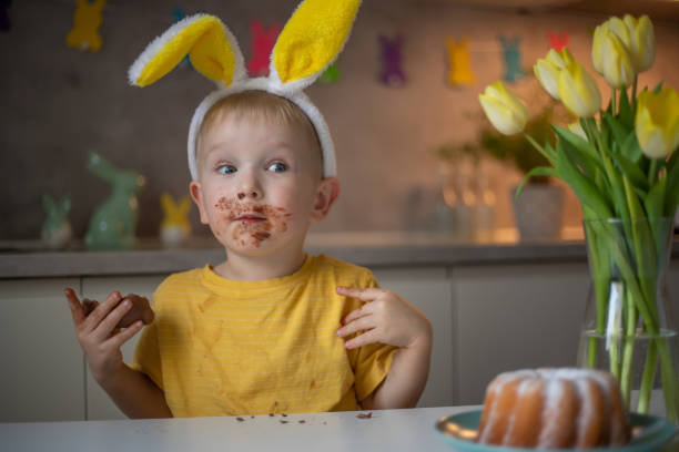A cute little boy wearing bunny ears on Easter day is eating a chocolate Easter bunny. A child plays egg hunt for Easter. Charming child celebrates Easter at home