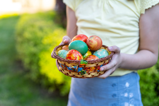 Close up of a girls hands holding decorated Easter eggs in a basket while standing outside in a garden