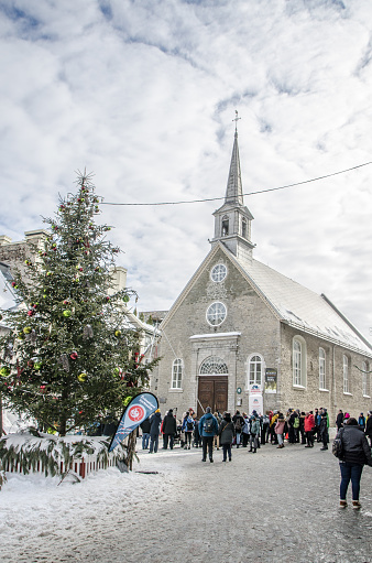 Crowd in front of church in Petit Champlain street during winter day in Quebec city.\nThere is a decorated Christmas tree with some winter Carnaval decoration