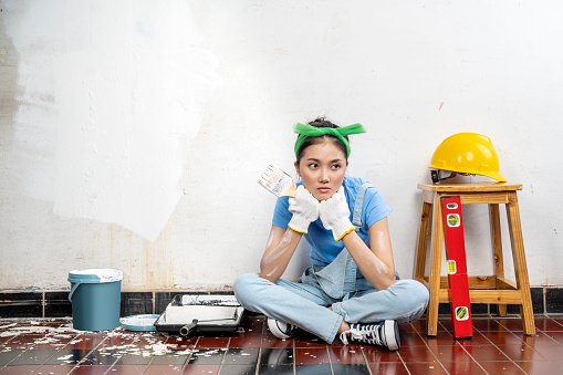 Asian young housewife in a glove holding a paintbrush with an unhappy expression with white wall background