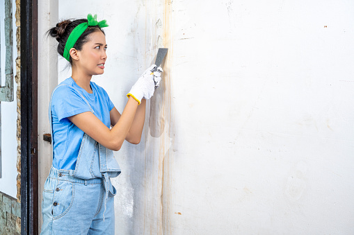 Asian young housewife in glove using a paint scraper on the wall