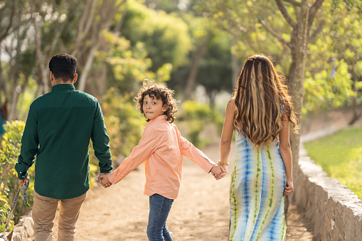 Kid turning to look at camera while walking holding hands with his parents in a park