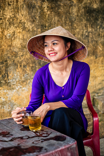 Vietnamese woman drinking traditional iced coffee - cafe sua da in old town in Hoi An city, Vietnam. Cà phê sữa đá is Vietnamese iced coffee with sweetened condensed milk. Hoi An is situated on the east coast of Vietnam. Its old town is a UNESCO World Heritage Site because of its historical buildings.