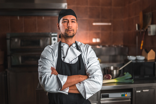 Front portrait of a good looking japanese chef standing in the kitchen. He is wearing a white uniform and has his arms crossed.