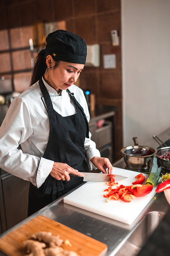 Waist up shot of female chef working in the kitchen. She is cutting a red pepper into small pieces for a recipe.