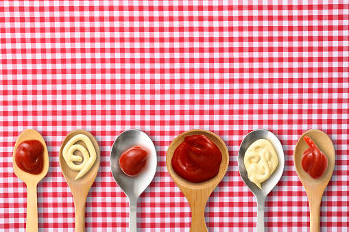 Overhead shot of Ketchup and mayonnaise on lined spoons on gingham checked tablecloth.