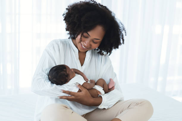 美しいアフリカ系アメリカ人の若い成人の母親がベッドに座り、生まれたばかりの赤ん坊を腕に抱き、笑顔で彼を見下ろしている。幸せな黒人家族素敵な、保育園の母乳育児母の日。 - breastfeeding mother newborn baby ストックフォトと画像