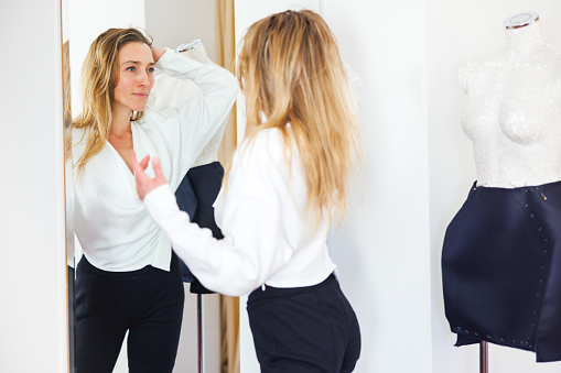 Woman wearing a newly designed white shirt, looking at her own reflection in the mirror with hand in hair, feeling proud of her work