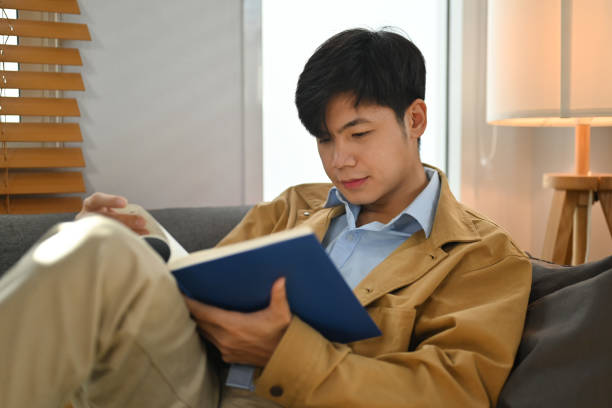 istock Calm asian man sitting on comfortable sofa and reading interesting book, enjoying leisure weekend time at home. 1465469647