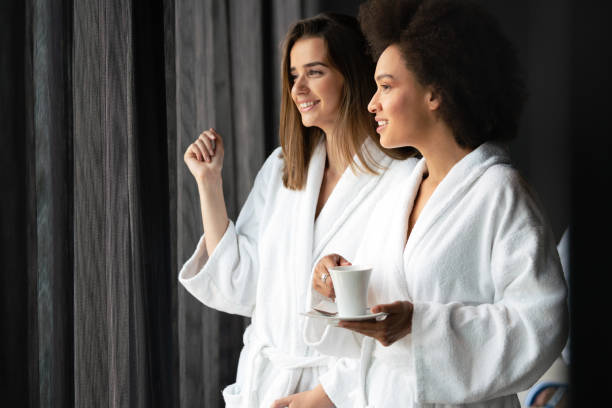 pareja de lesbianas relajándose y bebiendo té en batas durante el fin de semana de bienestar - bathrobe women cup towel fotografías e imágenes de stock