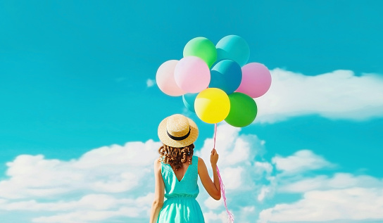 Rear view of young woman with bunch of colorful balloons wearing summer straw hat on the field on blue sky background