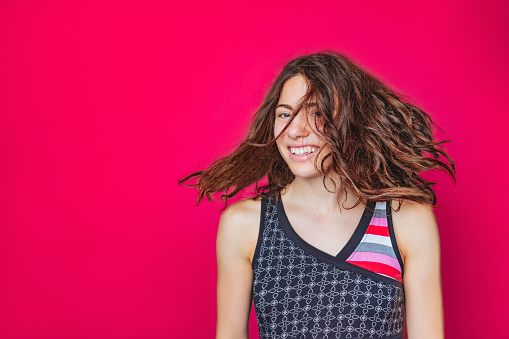 Portrait of beautiful young woman wearing colorful shirt, tossing her brown wavy hair and smiling cheerfully, magenta background, studio shot