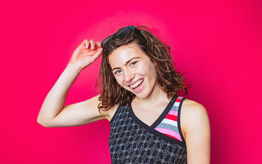 Portrait of beautiful young woman putting sunglasses in brown wavy hair, wearing colorful shirt and smiling joyfully at camera, magenta background, studio shot