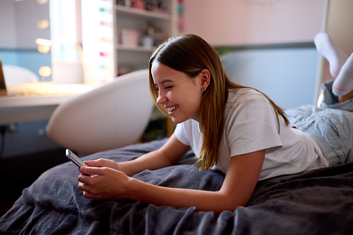 Smiling Teenage Girl Lying On Bed At Home Looking At Mobile Phone