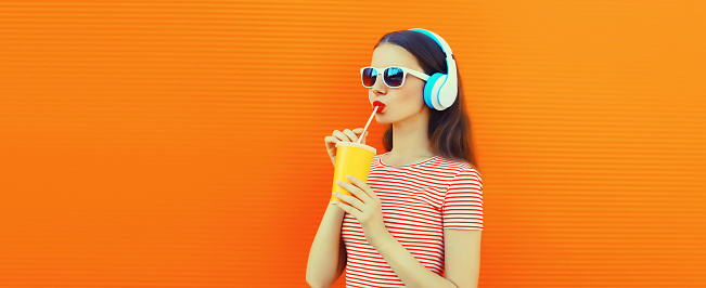 Portrait of young woman in headphones listening to music drinking fresh juice on orange background