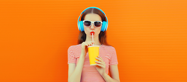 Portrait of young woman in headphones listening to music drinking fresh juice on orange background
