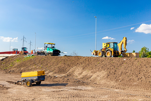 Zrenjanin, Vojvodina, Serbia - June 8, 2021:Spreader machine lays, pours hot asphalt, laying asphalt. Pneumatic compressor and excavator parked on construction zone. Road works on unfinished bridge.