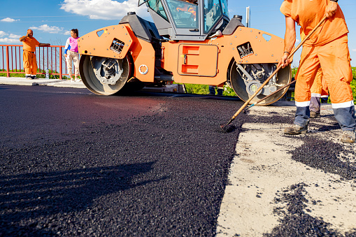 Zrenjanin, Vojvodina, Serbia - June 8, 2021: Workers are using rakes to level, set up layer of fresh tarmac to right measures, steamroller is flatting fresh asphalt .