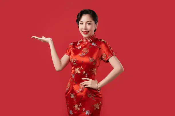 Happy Chinese new year, Beautiful young Asian woman wearing traditional cheongsam qipao dress with gesture of introduce isolated on red background,
