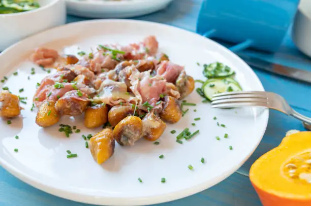 Delicious and easy gluten free dinner or lunch with pan fried pumkin, potato gnocchi, italian ham, melted cheese and herbs. Served ready to eat on a plate on blue wooden table background. Closeup