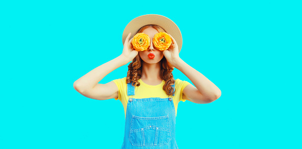 Summer portrait of happy young woman covering her eyes with flowers as binoculars looking for something wearing round straw hat, jumpsuit on blue background