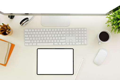Top view of a modern white office desk with digital tablet white screen mockup, keyboard, computer, wireless mouse, a coffee cup and stationery.