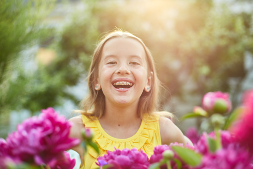 Cute baby girl 5-6 year old holding rose flower sitting over blooming bushes in garden outdoor. Spring season. Stylish little child over nature background. Springtime. Childhood.