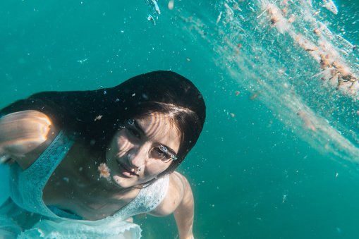 A young woman will dive with a white dress swimming, underwater image, summer lifestyle of a young Caucasian brunette looking at camera