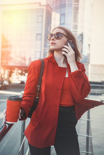 Young woman with a cup of coffee in her hands with makeup and glasses looking to the side while standing near her office. Portrait of a business girl near a glass building